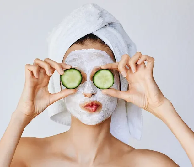 Kvinna med handduk på huvudet och ansiktsmask, hon håller två grukskivor framför ögonen.