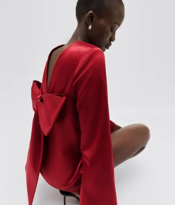 modell som sitter ner i en röd, kort klänning med v-ringad rygg och rosettdetalj.