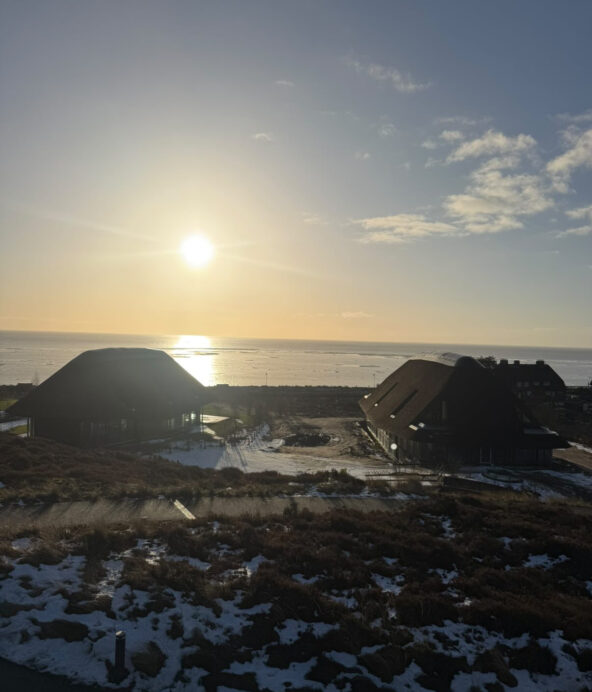 Utsikt över två hus ner vid vattnet samt sandstranden i solnedgång.