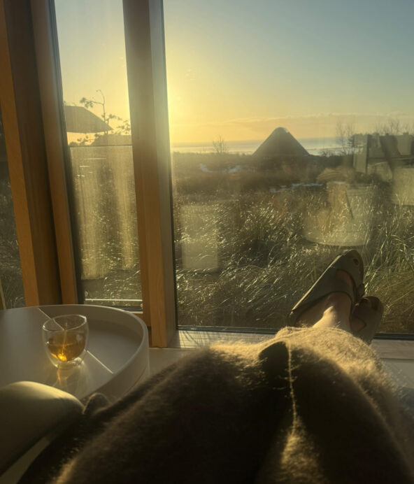 Fötter mot immigt fönster och utsikt över hus, stranden och havet. bredvid på ett bord står ett glas varmt the.