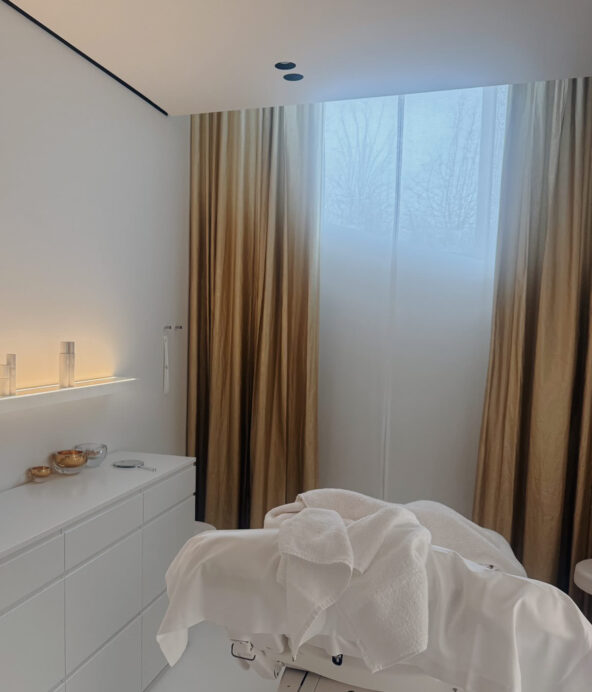 Behandlingsrum på Lanserhof hälsocenter. Beiga gardiner, vita handdukar, vita möbler och tända jus för e lugn och harmonisk känsla.