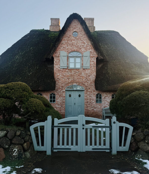 Ett fint gammalt hus i tegel omringat av fluffiga buskar. För att komma in till huset får man gå igenom en ljusgrön grind i trä.