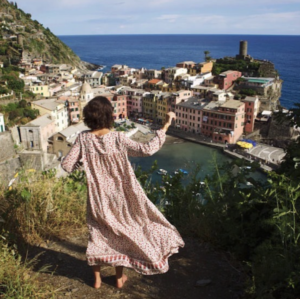 En kvinna med kort, lockigt, mörkt hår som dansar framför en utsikt över en italiens by. Kvinnan står ovanför byn på en klippa i en vid, rosa långklänning.