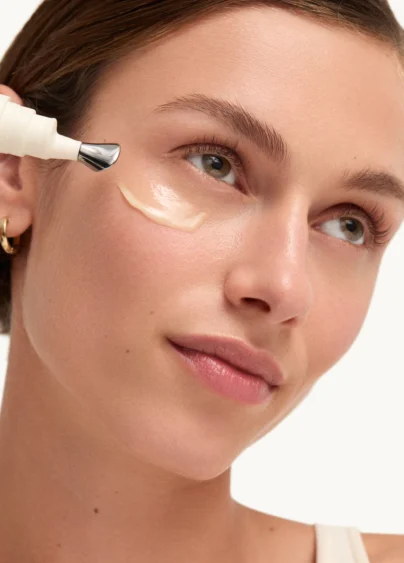 Kvinna som applicerat ögonkräm från Goop under ögonen. Hon har en swoosh av kräm under ögat och tittar uppåt och ler.