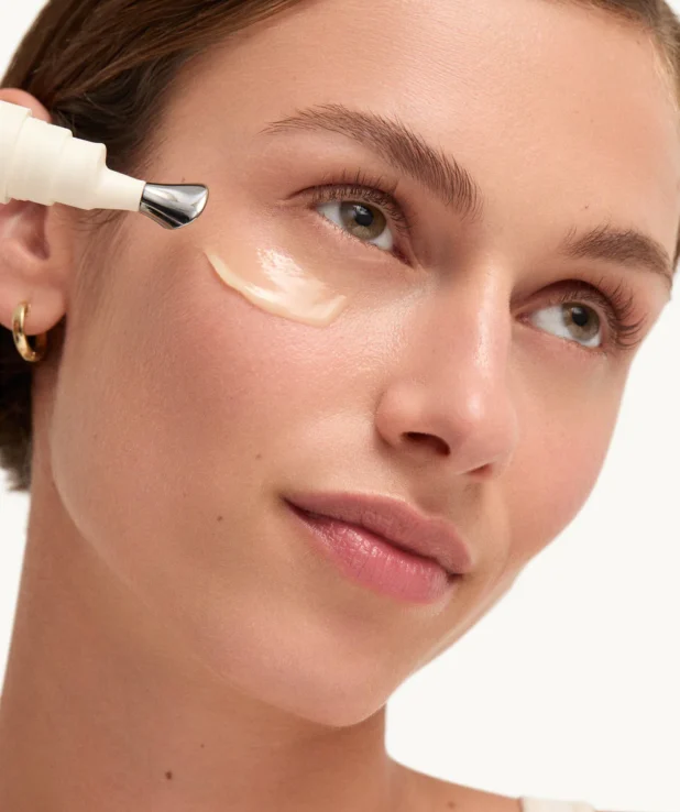 Kvinna som applicerat ögonkräm från Goop under ögonen. Hon har en swoosh av kräm under ögat och tittar uppåt och ler.