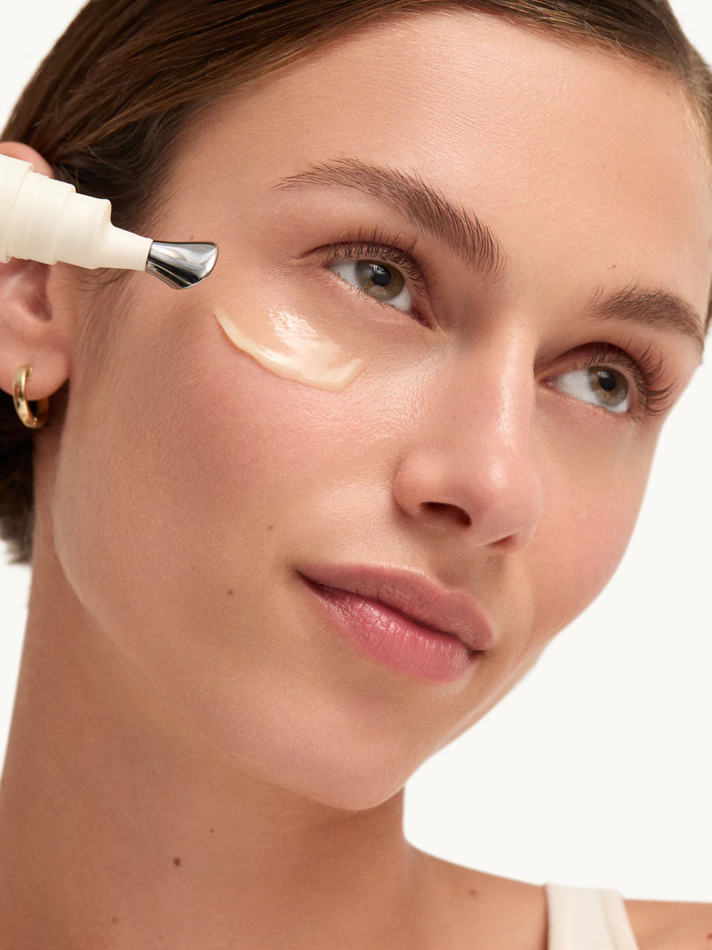 Kvinna som applicerat ögonkräm från Goop under ögonen. Hon har en swoosh av kräm under ögat och tittar uppåt och ler.