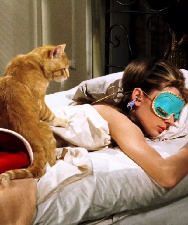 Audrey Hepburn från filmen "Breakfast in Tiffany's". Hon ligger i sängen med sovmask över ögonen och hennes katt sitter bredvid på sängen och tittar när hon sover.
